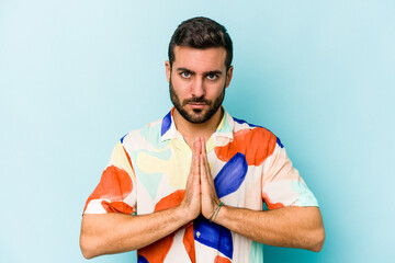 Young caucasian man isolated on blue background praying, showing devotion, religious person looking for divine inspiration.
