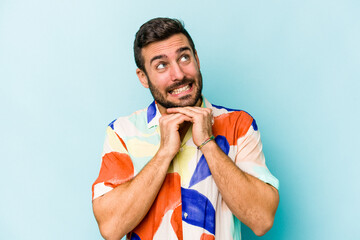 Young caucasian man isolated on blue background keeps hands under chin, is looking happily aside.