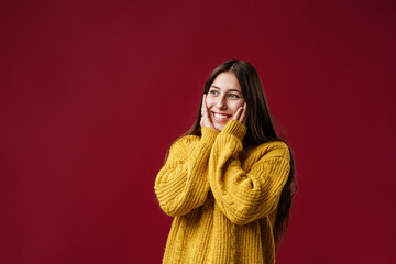 Portrait of a happy girl with dark long hair, smiling in a yellow knitted sweater. A teenager, emotionally excited, looks into the camera, standing on a red background in the studio