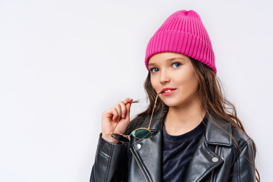 stylish teenage girl with dark hair, dressed in fashionable clothes, a leather jacket and a pink hat and glasses on white wall and looking at camera. transitional age. concept of youth and clothing.