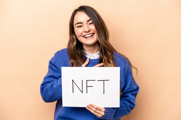 Young caucasian woman holding a NFT placard isolated on beige background laughs out loudly keeping hand on chest.