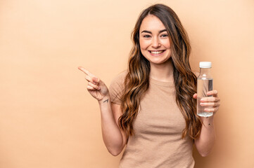 Young caucasian woman holding a bottle of water isolated on beige background smiling and pointing aside, showing something at blank space.