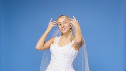 Fototapeta na wymiar Young beautiful blonde smiling in a wedding white dress and veil on a blue background. The girl looks into the camera. Wedding