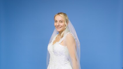 Fototapeta na wymiar Young beautiful blonde smiling in a wedding white dress and veil on a blue background. The girl looks into the camera. Wedding