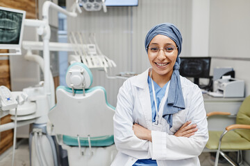 Waist up portrait of young female dentist looking at camera and smiling while wearing headscarf in dental office