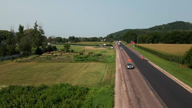 Drone view over rural highway being repaired with new asphalt. Pilot car leading traffic through the construction zone. Lush green landscape and farm field in the valley with forest covered mountains.