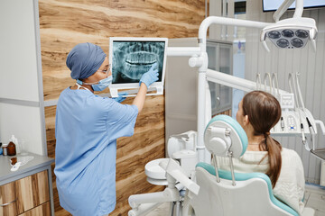 Back view of female dentist pointing at teeth X-ray while consulting patient in clinic