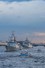 small boats with tourists inspect warships on the Neva in the city center against the blue evening sky