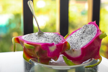 A sliced dragon fruit with a spoon stuck into the white sweet flesh against a background of greenery. Exotic tropical fruit