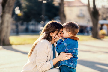 A little boy kissing his mommy while she hugging him in a park.