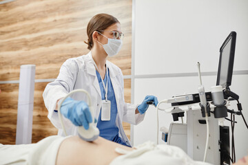 Side view portrait of female doctor using ultrasound machine and wearing mask while examining...