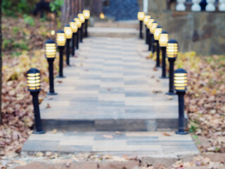 Low street lights are located along a tiled walkway with steps in the open air, surrounded by autumn foliage. Blurred image