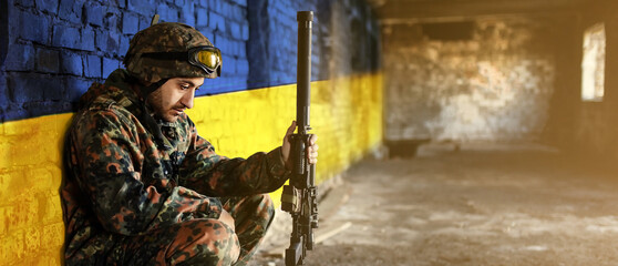Military soldier on the background of the flag of Ukraine. The flag of Ukraine is painted on a brick wall, a tired sad soldier sits with a weapon in his hands. Relations between Ukraine and Russia - Powered by Adobe