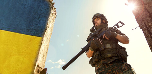 Ukrainian Soldier military in the war with a weapon in his hands. The flag of Ukraine is painted on a brick wall. Relations between Ukraine and Russia