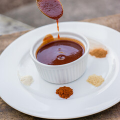 Still life of barbecue sauce in a small bowl with fresh spices on a white plate