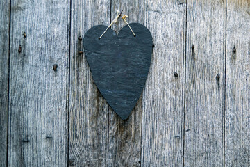 Plain black slate heart plate hung on a wooden surface with rusted nails, flat rock heart shaped sculpture on rustic wood. Blank space for copy.