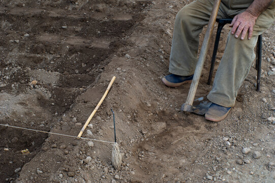 Pies de persona mayor agricultor sentada descansando, con azada en el huerto