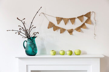 A green jug with cut poplar branches is on a white fireplace as a room decoration