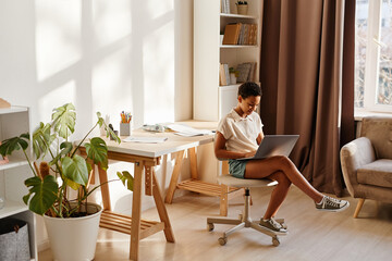 Minimal wide angle portrait of teenage girl using laptop at home in cozy interior lit by sunlight, copy space
