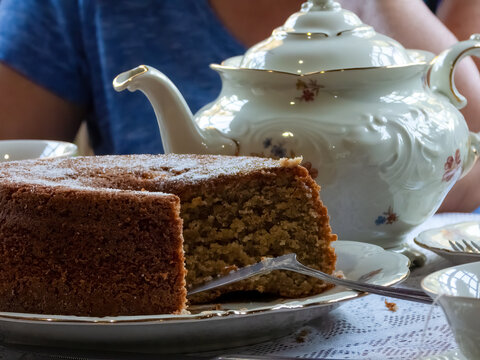 Cake cut on a table with white KPM porcelain tea set, decorared with flowers. White lace tablecloth, silver spatula, tea pot. Behind, a woman is sat (show hand and blue shirt).
