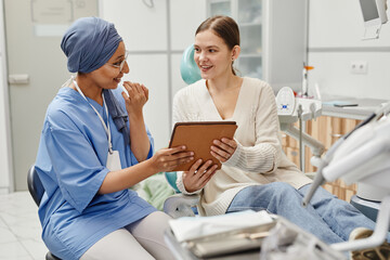 Portrait of smiling young woman in dental chair talking to female doctor and holding digital tablet
