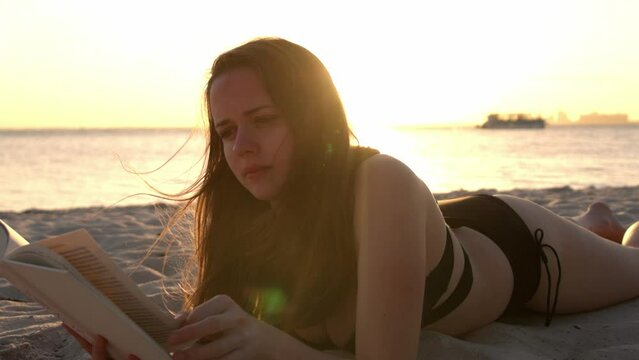 Young woman reads a book while lying on the beach at sunset