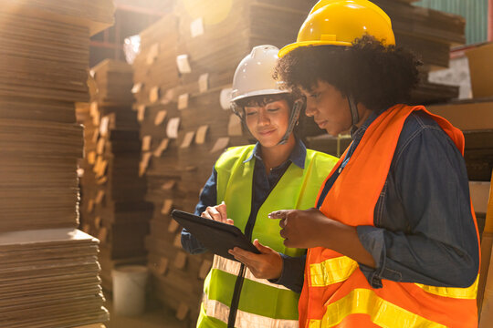 Engineer asian and african woman wearing safety helmet and vest holding clipboard and take note on the paper in the automotive part warehouse.Products and corrugated cardboard.