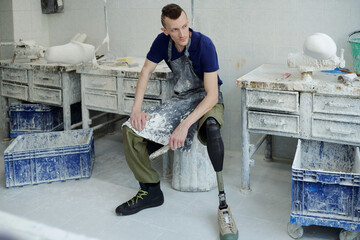 Young serious man with disability wearing apron sitting by workplace and having rest after work in...