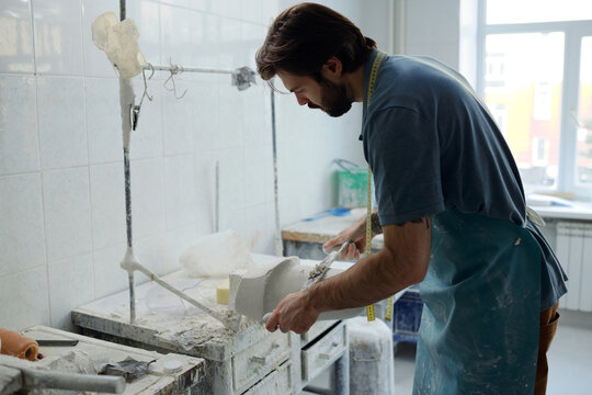 Young man bending over plaster cast while removing upper layer of material and smoothing its surface by workbench