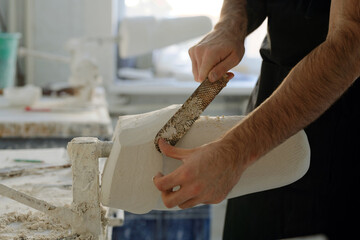 Hands of young factory worker with handtool removing excessive plaster from surface of cast while standing by workbench