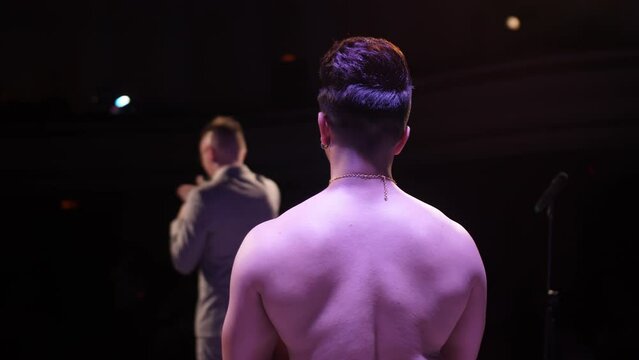 An Indian man without outerwear stands on stage. The bare back of an Indian dancer. Big stage. The host speaks on the microphone.