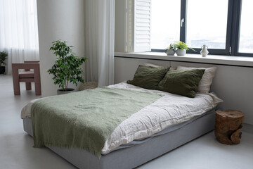 Double bed with cushions of white and treetop green colors and grey plaid standing by large window...