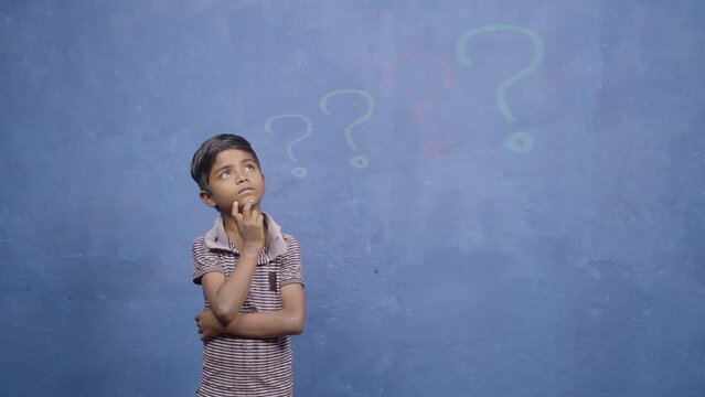 Poor Indian kid deep thinking on blue studio background with blinking big question marks - concept of thoughtful about future, wisdom and poverty.