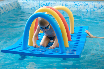 Toddler child crawling through tunnel on the water. Kid playing and learning to swim dive with equipment and toys in swimming pool in goggles. Boy doing early physicals development activities