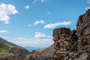 Scenic landscape with sunlit stone outliers with view to mountain vastness under cloudy sky. Colorful mountain scenery with sharp rocks on mountain pass under clouds in blue sky at changeable weather.