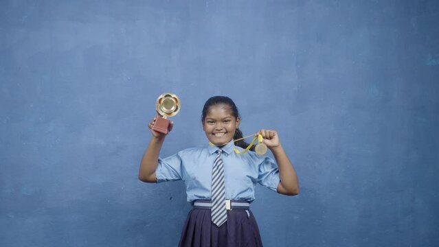 Enjoying indian school girl kid celebrating by showing trophy and medal by looking at camera - concept of achievement, proud, successful.