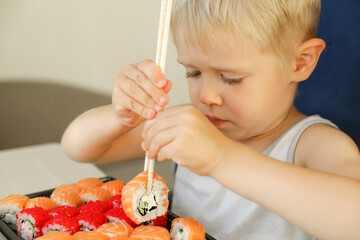 The child is eating sushi. Boy learning to eat philadelphia rolls with chopsticks.