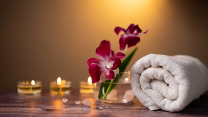 Fototapeta na wymiar Close up purple orchid flower, white clean rolled towel and candle over wooden table background. Facial beauty spa set, wellness well-being lifestyle concept.