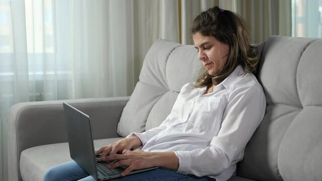 Young disabled woman with cerebral palsy works remotely on computer trying to type text by poorly mobile fingers sitting on sofa by window at home.