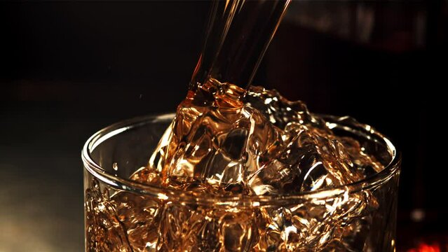 A jet of whiskey with splashes flows into a glass of ice. Against a dark background. Filmed is slow motion 1000 fps.