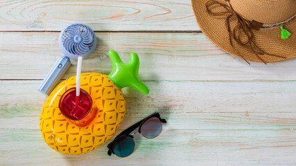 Closeup studio top view shot colorful pineapple shape rubber ring floating holder with iced cold drink glass with straw, sunglasses, small fan and weaving rattan hat with string hatband on wood table
