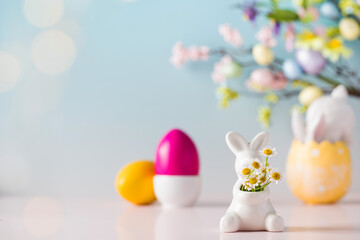Easter card. A white bunny with a bouquet of daisies on a blue background and colored eggs. Copy space.
