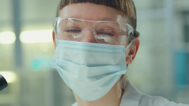 Portrait of young female scientist in sterile face mask and protective glasses posing on camera in laboratory