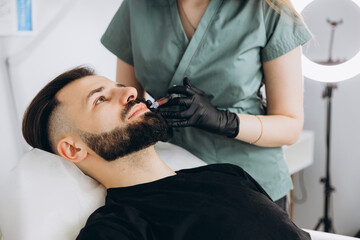 Fototapeta premium Stylish bearded man visiting aesthetic clinic, getting lips filler, closeup. Attractive Man having beauty injection at male spa salon. Anti-aging treatment for men concept.