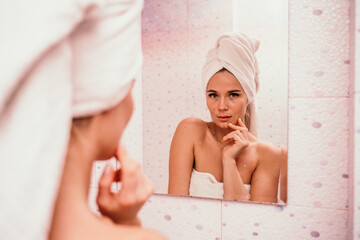 Portrait of young girl with towel on head in white bathroom looks and touches her face in the mirror and enjoys youth and hydration. Natural beauty, home care for problem skin