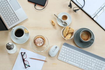 Meeting table seen from above with coffee cups and cookies, laptop, keyboard, folder and papers on a light wooden background, nobody
