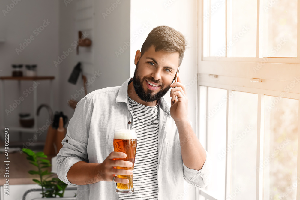 Wall mural Handsome bearded man with glass of beer talking by mobile phone in kitchen