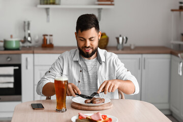 Fototapeta na wymiar Handsome bearded man eating sausages at table in kitchen