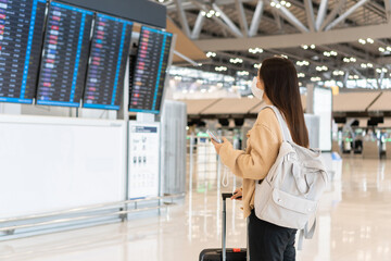 Young Asian woman wearing medical mask in international airport looking at the flight information board, checking her flight. Travel during coronavirus pandemic, new normal lifestyle concept.