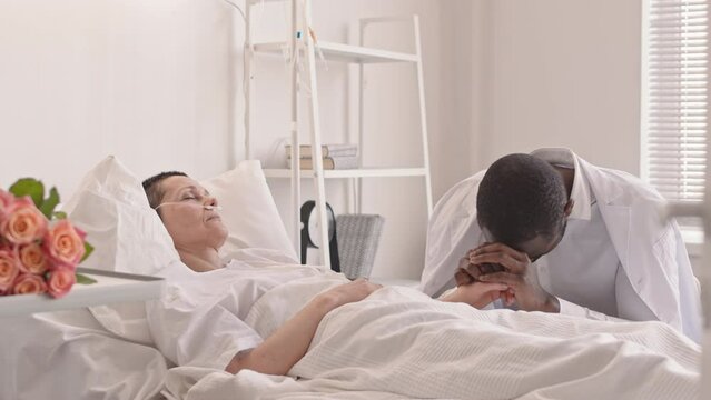 Medium shot of sad African American man holding hand of his sick wife in coma lying in hospital bed